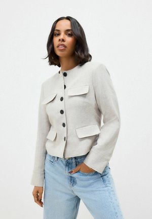 Hellgraue Leinenjacke mit rundem Ausschnitt, vorderen Knöpfen und zwei aufgesetzten Taschen. Die Ärmel sind leicht ausgestellt. Kombiniert mit hellblauen Jeans.
