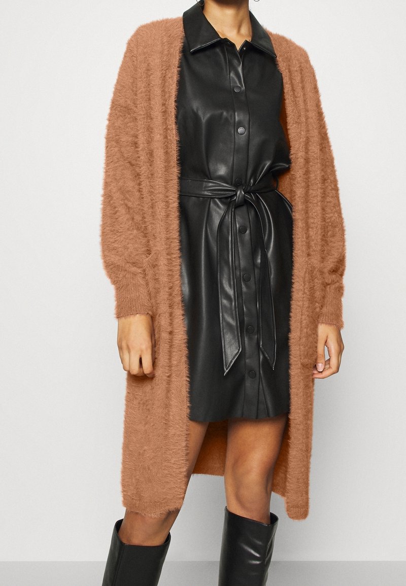 Femme portant une robe en cuir noire boutonnée avec une ceinture nouée à la taille, un long cardigan marron pelucheux, et des bottes noires montant jusqu'aux genoux.