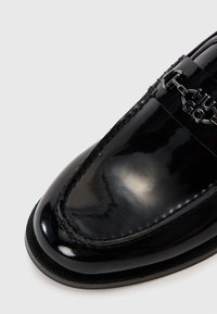 Mocasín de charol negro con un diseño elegante, que presenta una puntera cosida y un acento de logo metálico para mayor detalle. Textura suave y brillante.