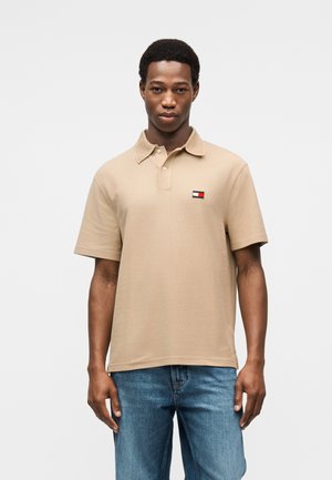 Beige polohemd van katoen met een kraag. Heeft een klein rechthoekig logo in rood, wit en blauw; gecombineerd met een blauwe spijkerbroek.
