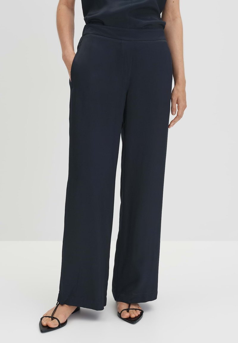 Marineblauwe wijde broek, gemaakt van soepel materiaal, met zijzakken en een elastische tailleband. De zoom heeft een subtiele splitdetail.