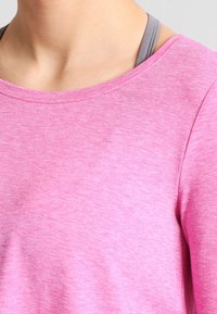 Haut long à manches en rose clair, avec un col rond. Tissu doux et texturé ; bretelles grises visibles d'un sous-vêtement en dessous.