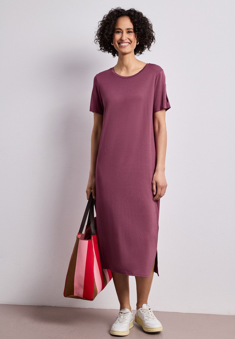Femme aux cheveux bouclés portant une robe midi violette, des baskets blanches et tenant un grand sac rayé, debout devant un fond uni.