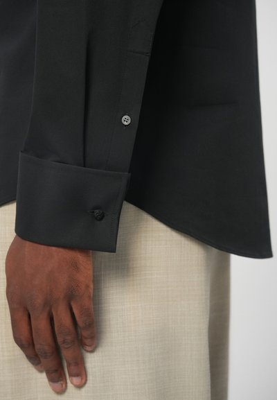 Μαύρο πουκάμισο με ομαλή υφή και μακριά μανίκια, που διαθέτει μανσέτα με κουμπί και διακριτές ραφές, συνδυασμένο με ελαφρύ μπεζ ύφασμα από κάτω.