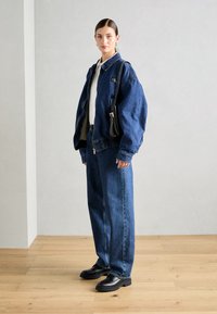 Veste en denim bleu foncé avec fermeture éclair à l'avant et poignets élastiques. Associée à un pantalon en denim bleu ample et des chaussures noires. Petit sac à main inclus.