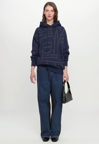 Navy hoodie with geometric line design, paired with wide-leg denim jeans featuring black side panels, and an embossed black handbag.