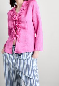 Blusa lunga a maniche con texture rosa e colletto arricciato, abbinata a pantaloni larghi a righe blu e bianche con tasche.