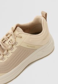 Baskets de sport beige clair avec tige en tissu doux, accents texturés et semelle rembourrée. La marque est visible sur le côté.