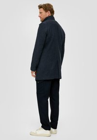 Marineblauwe gestructureerde jas met hoge kraag, enkele split en gestructureerde mouwen, gecombineerd met donkere broek en crèmekleurige sneakers.