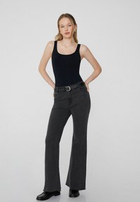 Mladá žena v černém bezrukávovém topu, vysokém černém džínovém pásku a černých botách, se zlatými kruhovými náušnicemi, stojící s rukama na bokách.