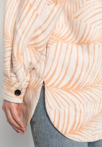 Camicia a maniche lunghe in morbido tessuto crema con un motivo di foglie corallo; presenta un orlo arrotondato e polsini con bottoni, indossata sopra un jeans chiaro.