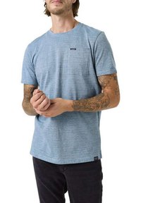 Camiseta de manga corta azul jaspeada con un bolsillo en el lado izquierdo del pecho; textura suave y cuello redondo; jeans oscuros visibles abajo.