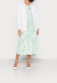 Bílý blejzr přes zelené a modré květinové šaty s naskládanou sukní. Modelka má na sobě bílé boty na blokovém podpatku a stojí na šedém pozadí.