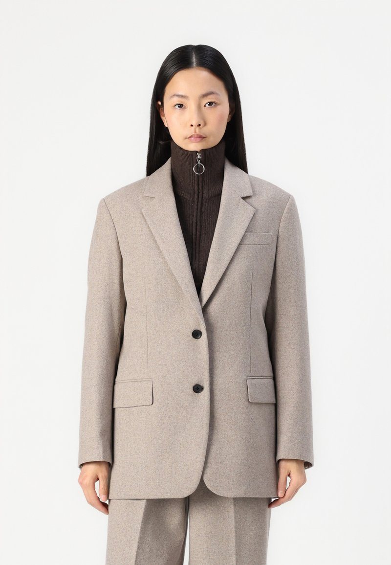 Blazer in lana beige con due bottoni neri, rever a risvolto e due tasche frontali. Collo a dolcevita marrone testurizzato sotto.