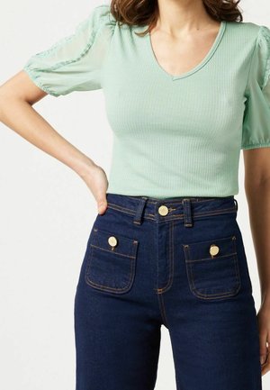 Top de canalé en menta claro con mangas de globo transparentes, combinado con pantalones cortos de mezclilla oscuros de cintura alta, que presentan detalles en botones dorados y bolsillos frontales.