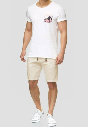 Wit T-shirt met korte mouwen en een roze palmboomprint op de zak, gecombineerd met beige shorts en witte sneakers. Casual, nauwsluitend ontwerp.