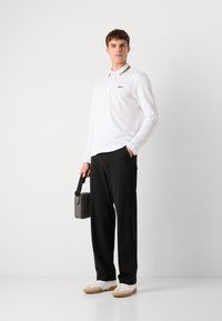 Langermet hvit polo skjorte med svart og gul krage, parret med svarte bukser og hvite joggesko. Grå veske i hånden.