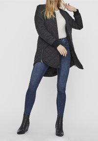 Giacca nera trapuntata con taglio lungo e dritto, polsini a coste e tasche laterali con zip, abbinata a jeans skinny blu e stivaletti neri.