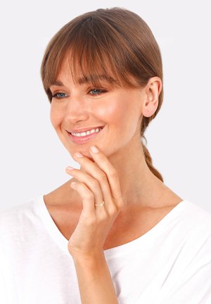 Goldring mit einem kleinen grünen Akzent, der an einer Hand getragen wird, mit einem schlichten Design. Der Hintergrund ist hellgrau, und die Person trägt ein einfaches weißes Oberteil.