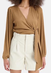 Blusa avvolgente marrone a coste con maniche lunghe, abbinata a pantaloni corti bianchi plissettati a vita alta. Il tessuto appare strutturato e morbido.