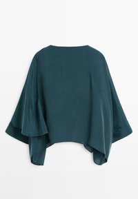 Långärmad, mörk teal-plagg med en svävande siluett, tillverkat av ett slätt tyg. Har vida ärmar och en rundad halsringning.