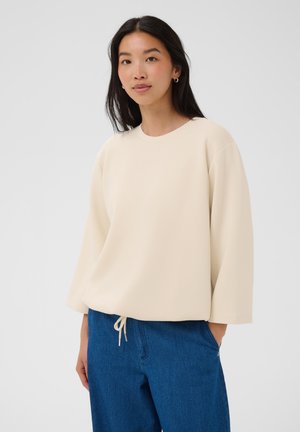 Frau mit langem dunklem Haar trägt einen lockeren cremefarbenen Pullover und blaue Jeans, steht mit einer Hand in der Tasche vor einem schlichten Hintergrund.