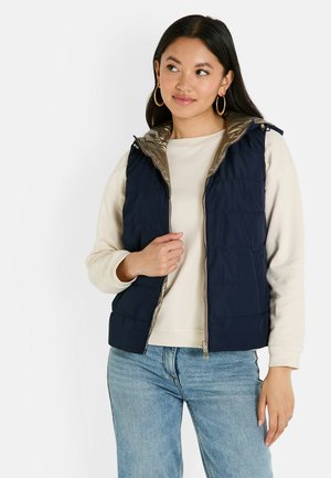Gilet matelassé bleu marine avec un intérieur doré brillant, doté d'un zip sur le devant, d'une capuche et de coutures verticales. Porté par-dessus un haut beige clair.