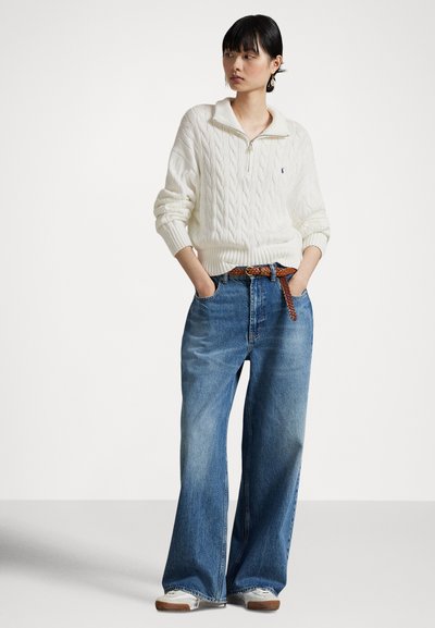 Γυναίκα που φοράει ένα λευκό πουλόβερ με φερμουάρ μέχρι τη μέση και πλεκτό σχέδιο, φαρδιά μπλε τζιν με καφέ πλεκτή ζώνη και λευκά αθλητικά παπούτσια, στέκεται με τα χέρια στις τσέπες.