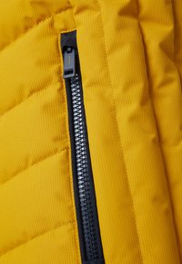 Gelber Pufferjackenstoff mit Kanälen und Steppnähten, ausgestattet mit einem schwarzen Reißverschluss und einer kontrastierenden Reißverschlussleiste entlang der Seitennaht.