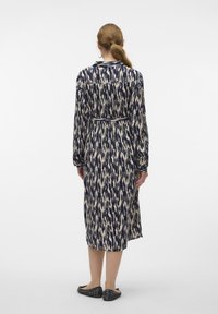 Långärmad klänning i marinblå och krämfärgad mönstrad tyg, knäppning fram, bälte i midjan och nedanför knäet längd. Bärs med platta skor.