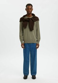 Chemise longue à manches longues couleur olive clair, pull marron drapé autour du cou, associée à un jean bleu à jambes larges et des chaussures noires. Design simple, textures douces.