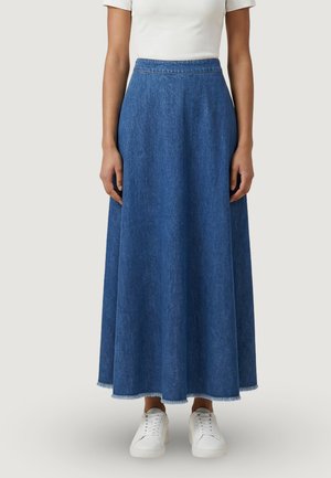 Žena v modré džínové sukni s vysokým pasem a délkou sahající k kotníkům, s neupraveným lemem a bílými teniskami, doplněná bílým topem s krátkými rukávy.