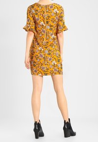 Žena v krátkém rukávu, hořčicově žlutých květovaných šatech se zipem na zadní straně, doplněná černými kotníkovými botami na blokovém podpatku.