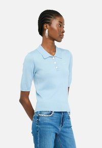 Polo-shirt bleu clair à manches courtes avec une finition texturée, col classique, patte de boutonnage à trois boutons et logo brodé. Porté avec un jean orné.