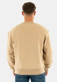 Man med kort brunt hår som bär en beige sweatshirt och blåjeans, stående med ryggen mot en neutral vit bakgrund.
