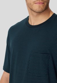 Camiseta de manga corta en teal oscuro con un estampado, cuello redondo y un bolsillo en el pecho que presenta un pequeño diseño geométrico.