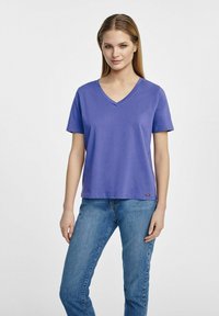 Fioletowy t-shirt z dekoltem w serek z miękkiego materiału, z krótkimi rękawami, w połączeniu z niebieskimi dżinsami. Posiada metkę na dołku dla identyfikacji marki.