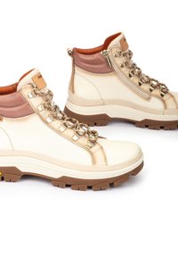 Botas de tobillo color crema con un acabado de cuero suave, acentos marrones contrastantes, ojales de metal y una suela de goma texturizada.