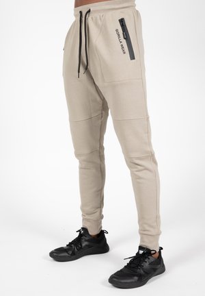 Beige jogger pants with a tapered fit, ribbed texture, and zippered side pockets. Features black drawstrings and a logo on the hip.