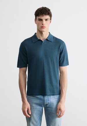 Jeune homme aux cheveux courts et bouclés portant un polo bleu à manches courtes et un jean bleu clair, debout devant un fond uni.