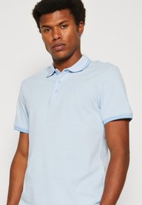 Polo de color azul claro, hecho de tela suave, con una tapeta de tres botones, logo bordado y ribete de contraste en las mangas.