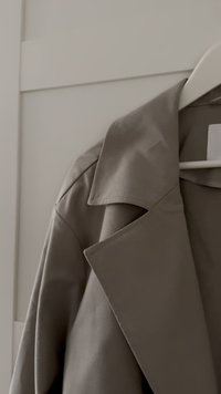 Svetlo sivi plašč s pasom, z oblikovanim krojem, velikimi ovratniki in gladko teksturo, obešen na belo obešalnik pred nevtralnim ozadjem.