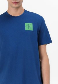 Tengerészkék pamut póló, amelyen egy négyzet alakú zöld logó található fekete "cK" felirattal. Rövid ujjú és kikerekített nyakkivágású, sima textúrával.