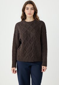 Brązowy sweter z dzianiny o teksturowanym wzorze diamentowym, okrągłym dekolcie i długich rękawach, zestawiony z ciemnoniebieskimi spodniami.