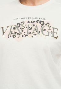 Λευκό πουκάμισο με το κείμενο "ΚΑΝΕ ΤΑ ΟΝΕΙΡΑ ΣΟΥ ΠΡΑΓΜΑΤΙΚΟΤΗΤΑ" πάνω από μεγάλα γράμματα "VINTAGE" σε ροζ και λεοπάρ φόντο.