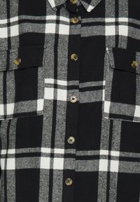 Camicia in flanella a quadri bianco e nero con chiusura a bottoni, due tasche sul petto e bottoni dorati. Texture morbida, motivo rettangolare.