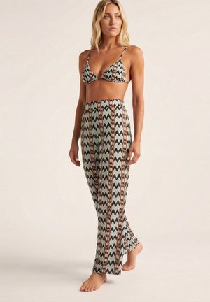 Mujer que lleva un conjunto de dos piezas a juego con top de bikini con patrón de zigzag en negro, marrón y verde y pantalones de cintura alta y pierna ancha.