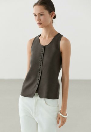 Vrouw die een mouwloze donkere top met knopen en hoge hals draagt en witte broek, staand tegen een effen lichte achtergrond.
