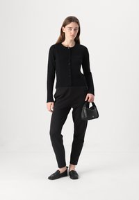 Cardigan nero con collo rotondo e chiusura a bottoni, abbinato a pantaloni neri a sigaretta, scarpe nere senza lacci e una piccola borsa nera.