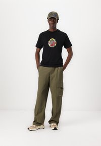 Black t-shirt with a colorful graphic design, olive cargo pants, and beige athletic shoes featuring white accents and a patterned cap.
